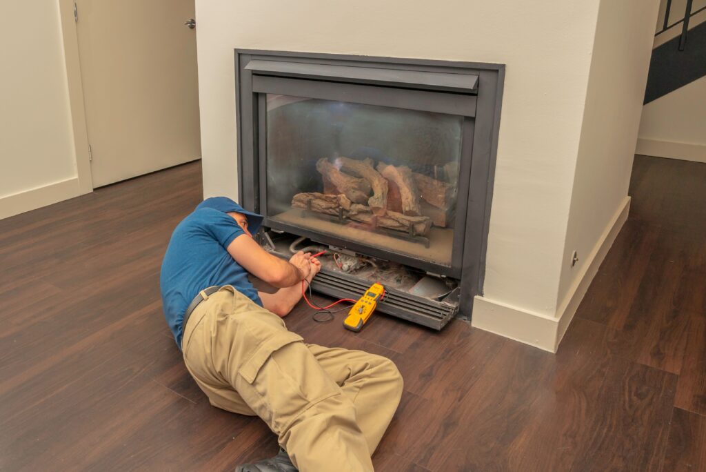 A technician in a blue shirt and beige pants lies on the floor, working on a fireplace with tools and a multimeter.